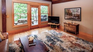 A spacious living room with a wooden floor, a large window, a television, a dining table, and a framed picture on the wall. - Free Night: Make Ozarks Memories at this Custom Lakefront Post & Beam Home VIDEO (Eldon)