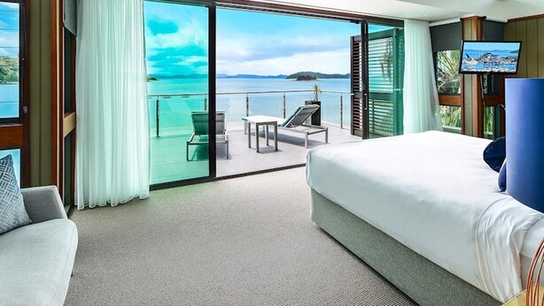 Una habitación de hotel moderna con una cama grande, un balcón con vistas al mar, un televisor y una zona de estar con una mesa y sillas.