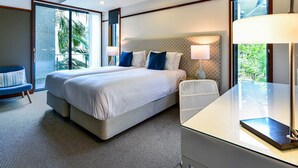 Habitación de hotel moderna con una cama grande, un área de descanso con una butaca, un escritorio y un ventanal con vistas a zonas verdes.