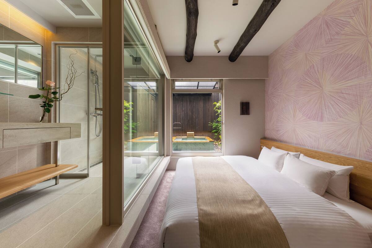 A modern bedroom with a large bed, a wooden bench, a glass shower enclosure, and a view of a courtyard.
