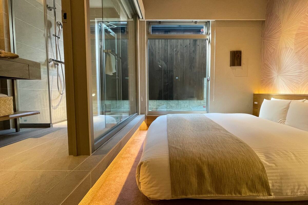 A modern hotel room with a large bed, a bench, a glass shower enclosure, and a view of the outdoors.