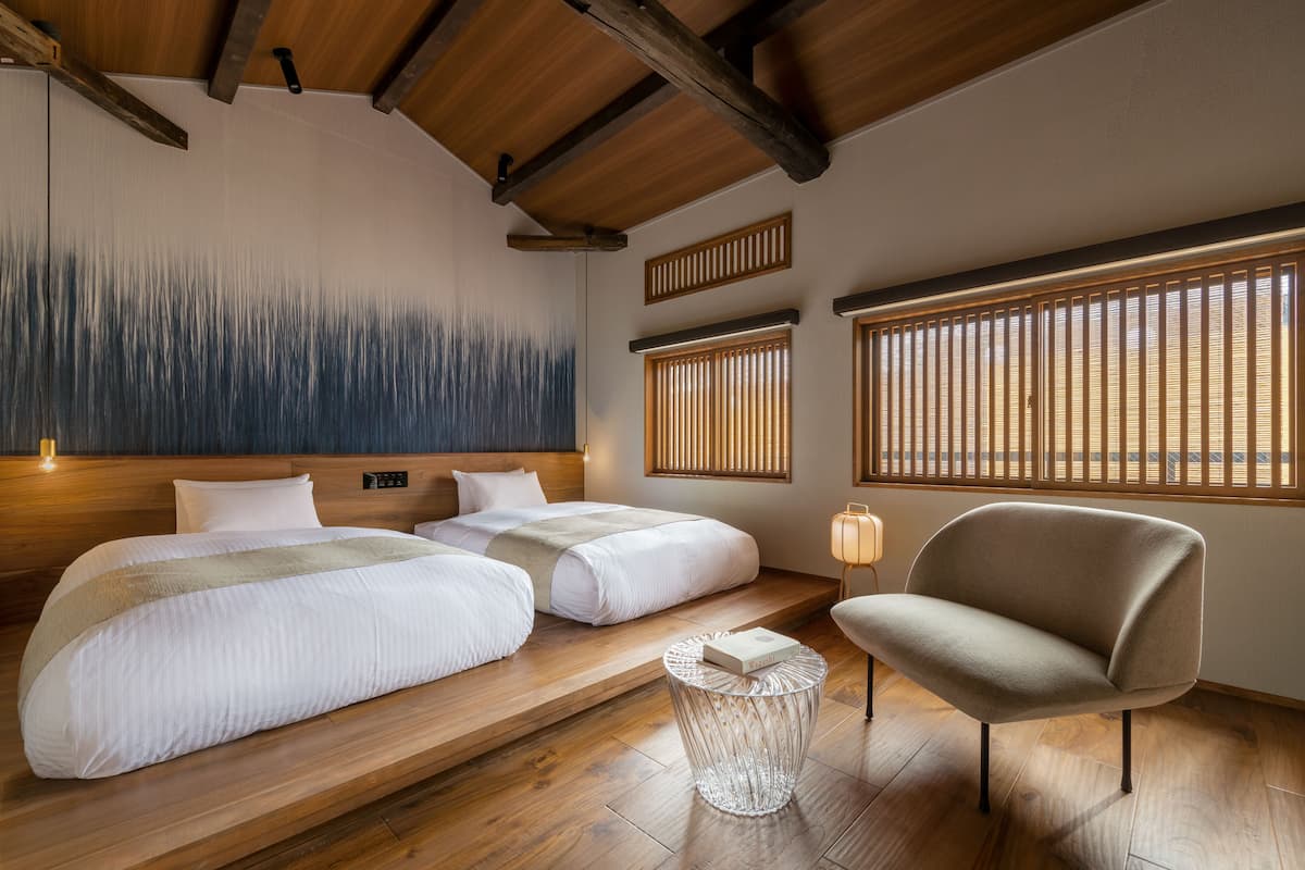 A hotel room with two beds, a chair, a small table, and a wall with vertical blinds.
