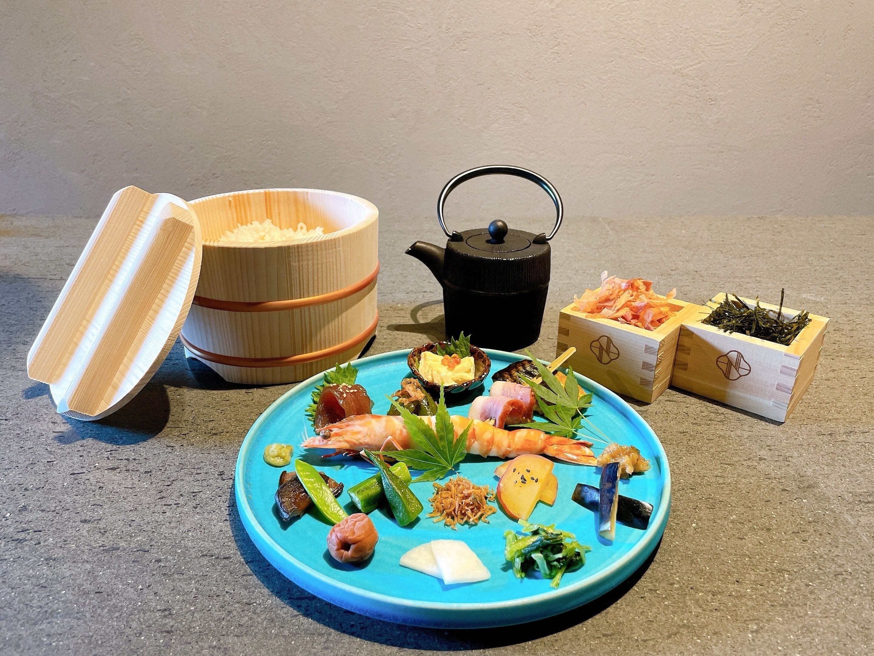 Een blauw bord met verschillende soorten eten, een zwarte theepot en houten dozen vol etenswaren.