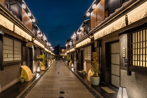 A narrow alleyway with traditional wooden buildings, potted plants, and illuminated signs.