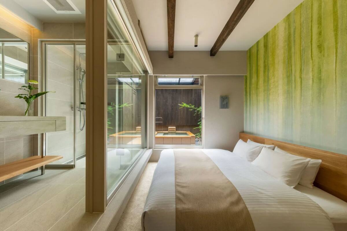 A modern hotel room with a large bed, a glass shower enclosure, a wooden bench, and a view of a pool and outdoor seating area.