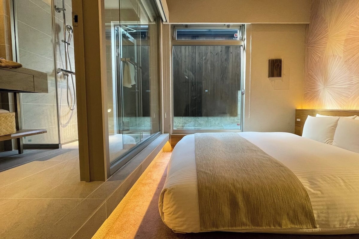 A modern hotel room with a large bed, a bench, and a glass door leading to a balcony.