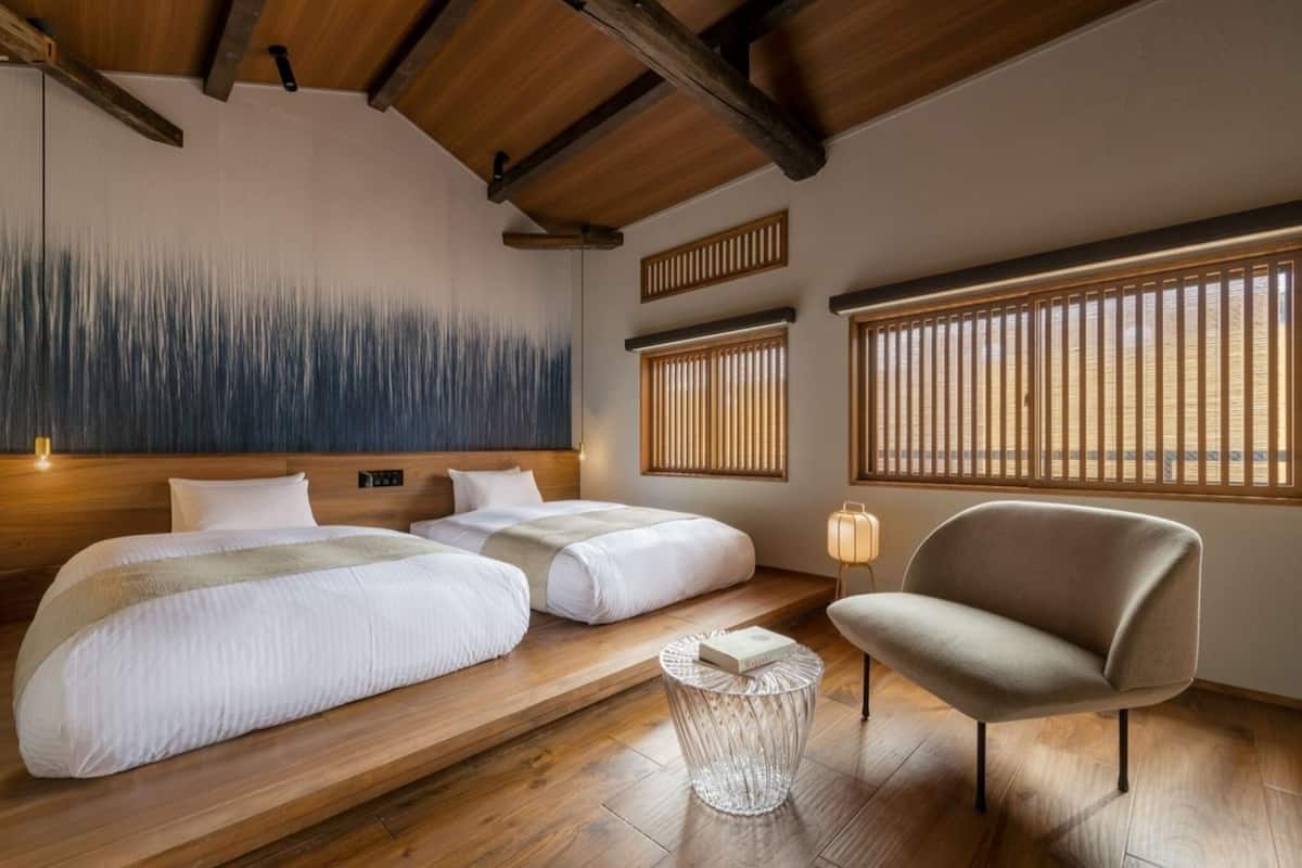 A hotel room with two beds, a wooden ceiling, a wall painting, and large windows with blinds.