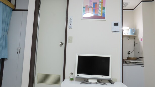 Une pièce avec une porte blanche, un meuble blanc sur lequel se trouve un téléviseur, une petite étagère avec des plantes et une affiche accrochée au mur.