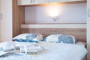 Ein ordentlich bezogenes Bett mit gemusterten Kissen und gefalteten Handtüchern, ein Holz-Kopfschild und eine wandmontierte Leuchte.