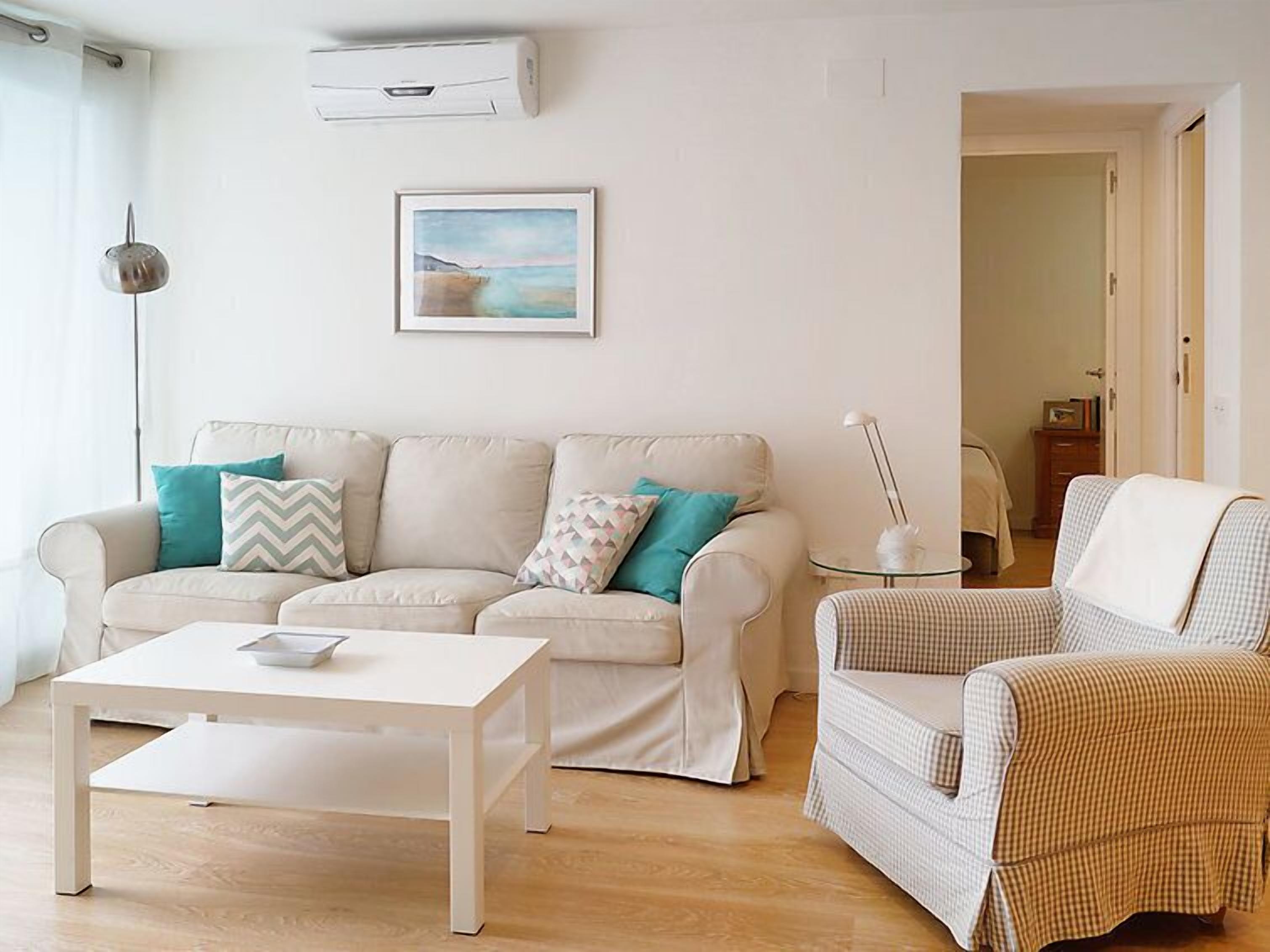 Ein Wohnzimmer mit einem beigen Sofa, einem weißen Couchtisch und einem karierten Sessel. An der Wand hängt ein Bild und über dem Sofa befindet sich eine Klimaanlage.