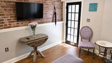 Ein Zimmer mit Backsteinwand, einem Flachbildfernseher über einem Holztisch mit Blumenvase, einer schwarzen Tür mit Glasfeldern und einem gemusterten Sessel.