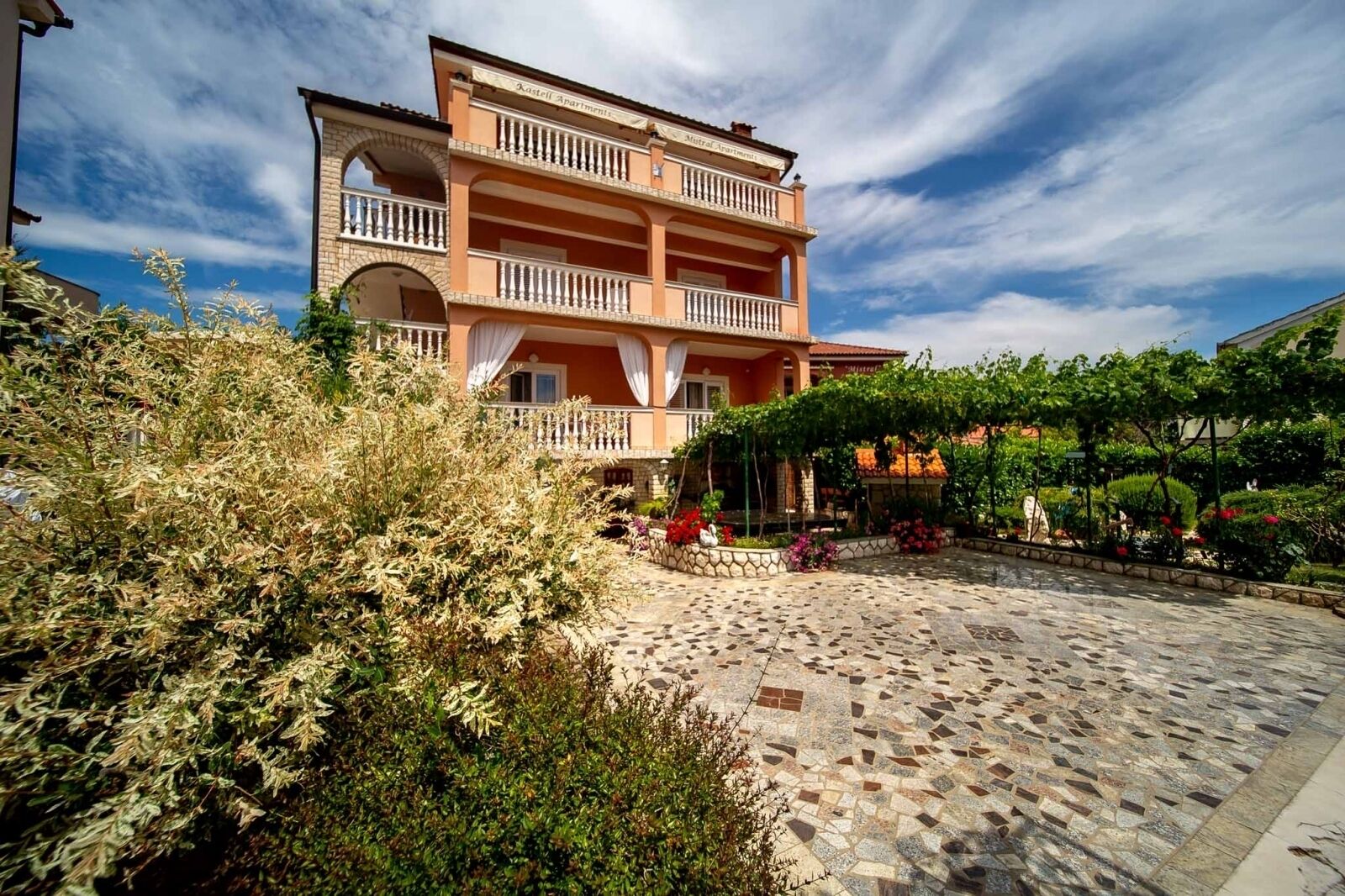 A two-story building with balconies, a courtyard with stone paving, and surrounding greenery.