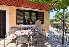 A covered outdoor dining area with a table set for two, a window with shutters, and a view of the garden.
