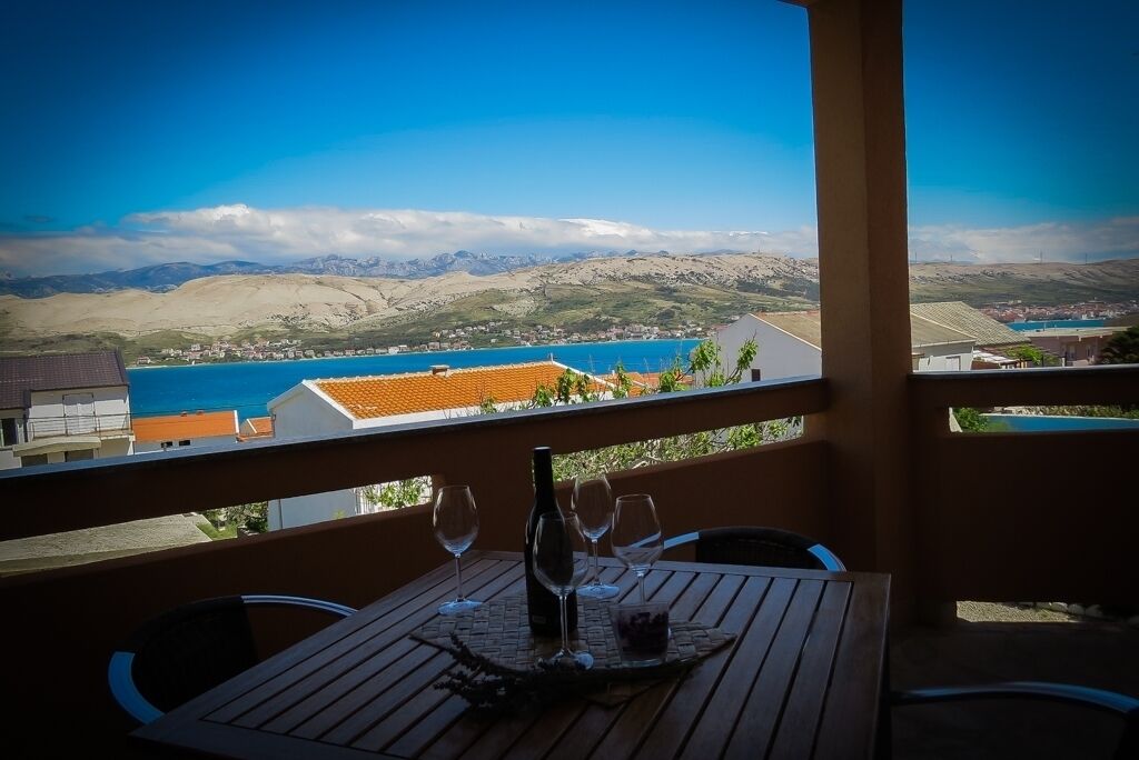A wooden table with four wine glasses and a bottle, overlooking a coastal town with mountains in the background.