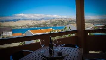 A wooden table with four wine glasses and a bottle, overlooking a coastal town with mountains in the background.