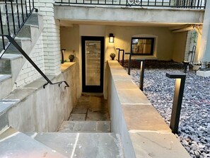 Eine Treppe führt zu einer Tür mit einem Fenster darüber, umgeben von einer Steinmauer und einem kleinen Garten mit Kies.