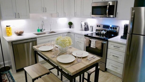 A modern kitchen with white cabinets, stainless steel appliances, a dining table set with plates and glasses, and a small plant on the counter. - No More Looking-Parking (Washington)