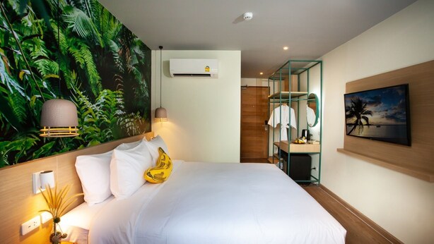 A hotel room with a bed, a wall-mounted air conditioner, a painting, and a bathroom with a shower and a towel rack. - The Stay Chaweng Beach Resort (Koh Samui)