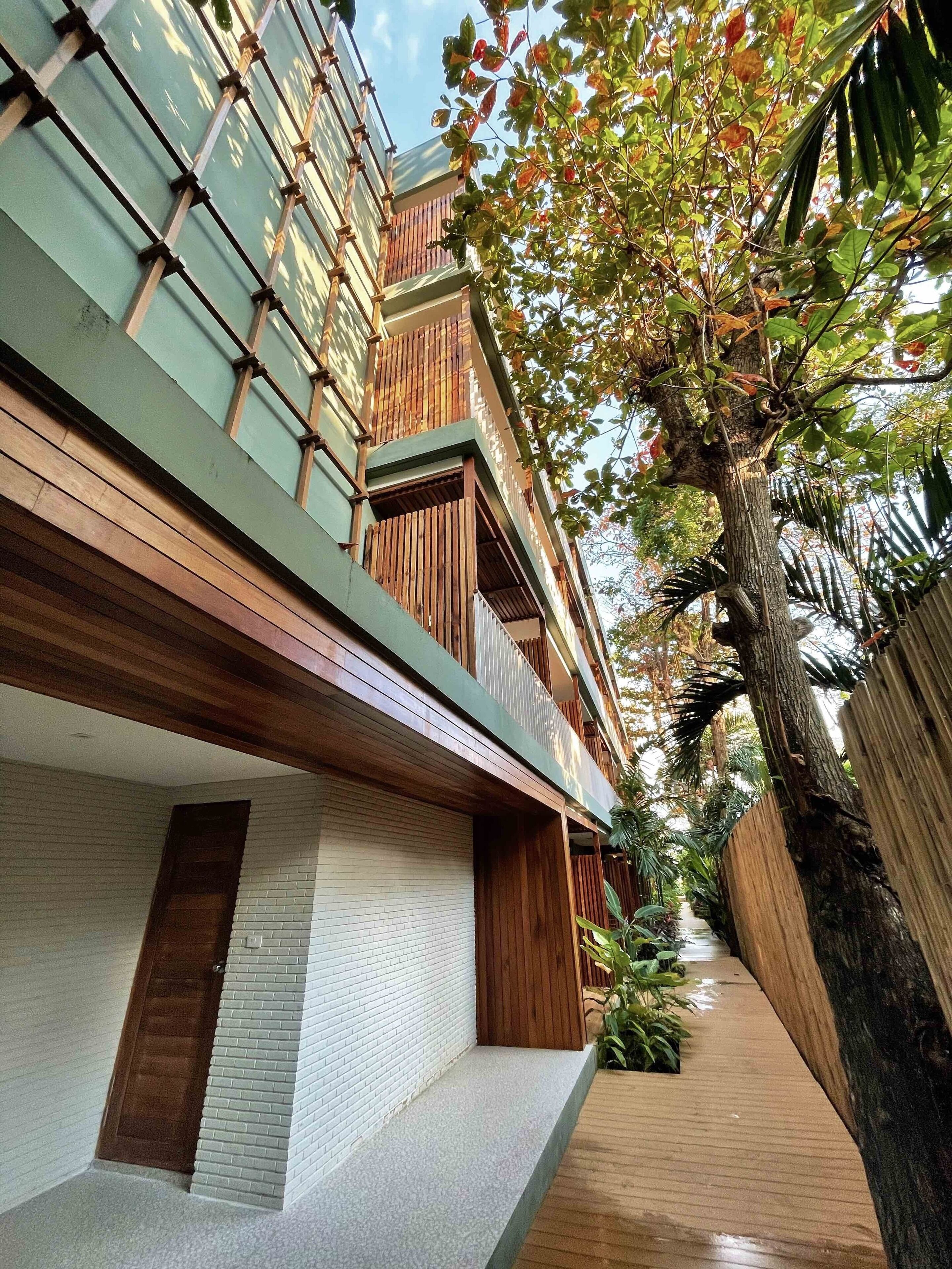 A modern building with a wooden balcony, a brick wall, and a tree-lined pathway.