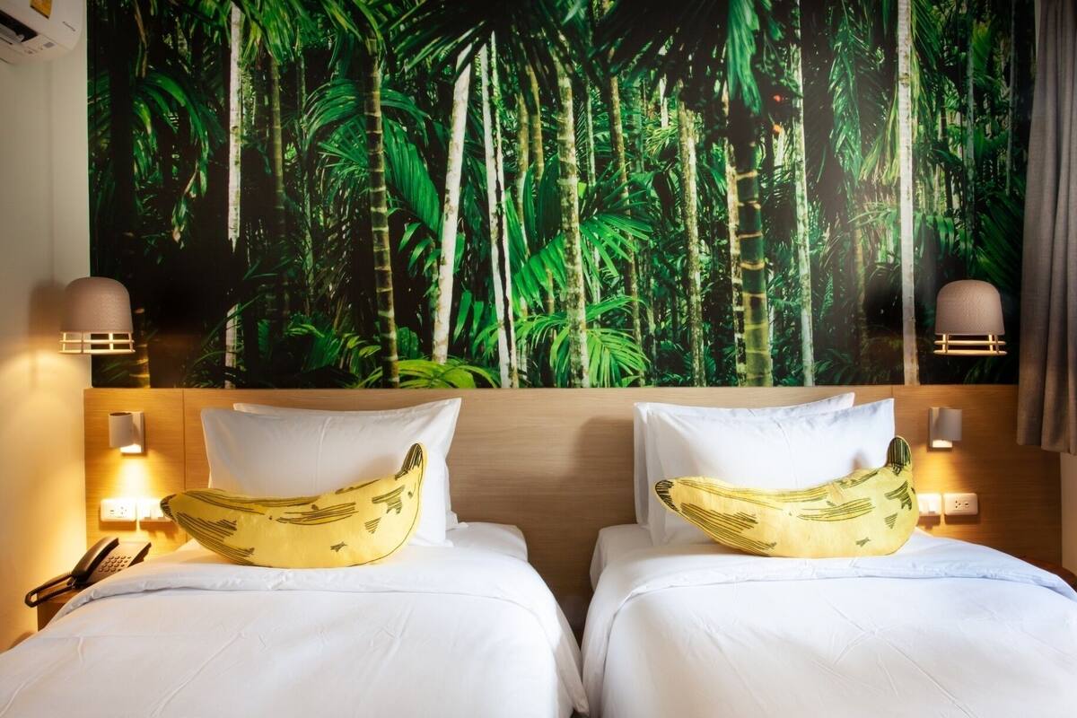 Quarto de hotel com duas camas, cada uma com colcha branca e travesseiro em formato de banana. Há um grande mural tropical na parede atrás das camas, e duas luminárias fixadas na parede, uma de cada lado das camas.