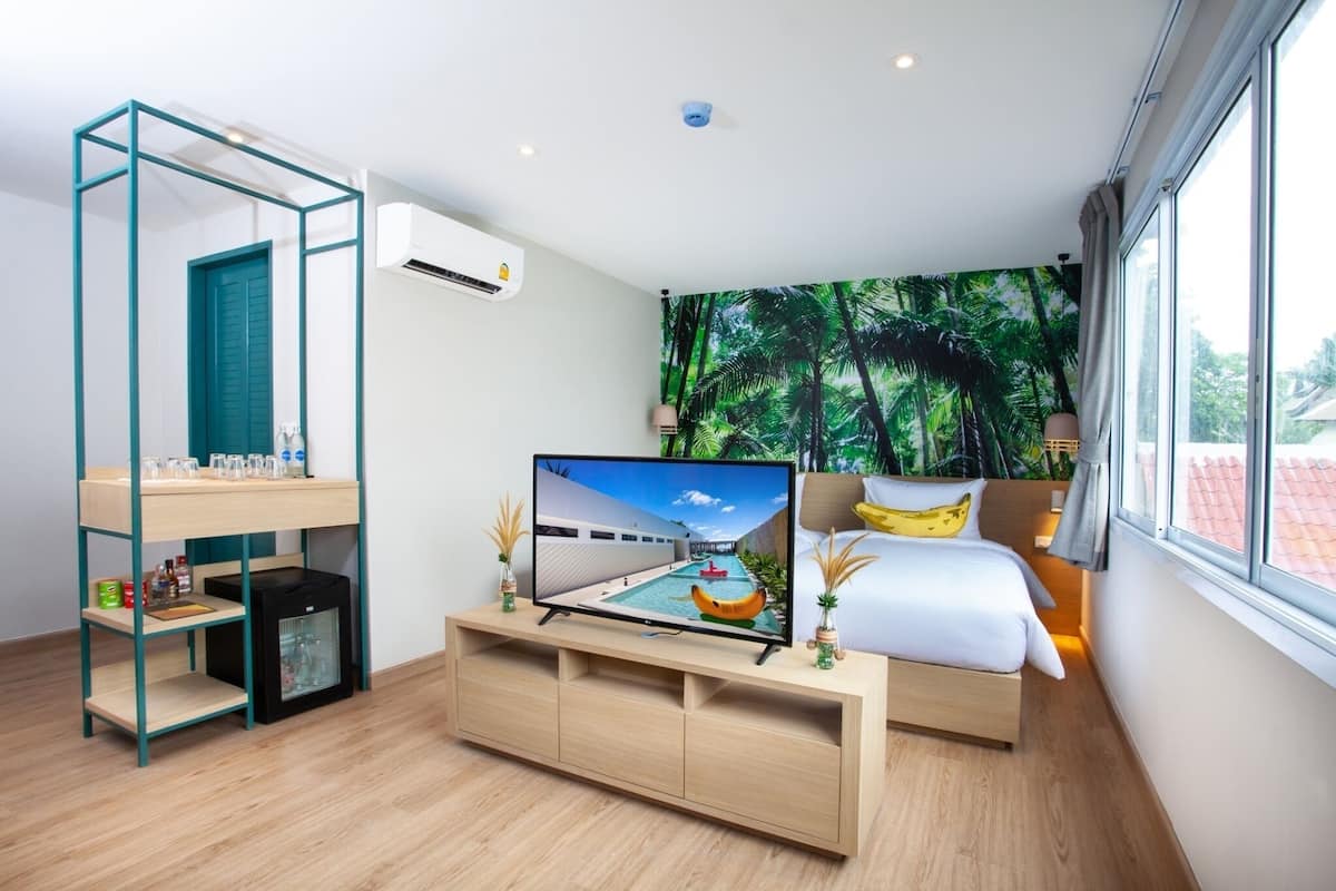 Quarto de hotel moderno com televisão, cama, escrivaninha e vista para palmeiras pela janela.