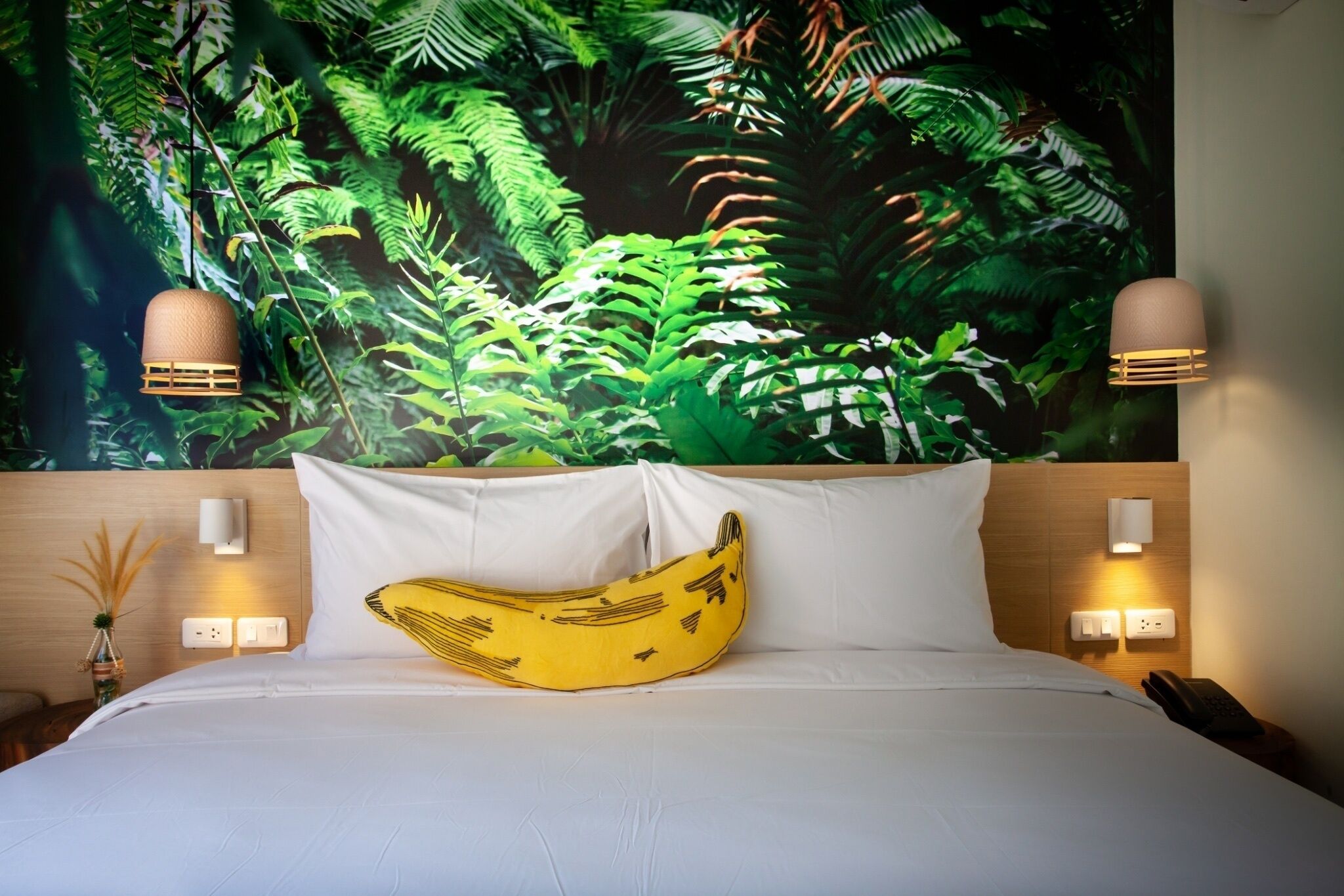 A hotel room with a bed, a banana-shaped pillow, a wall mural of green foliage, and two wall-mounted lamps.
