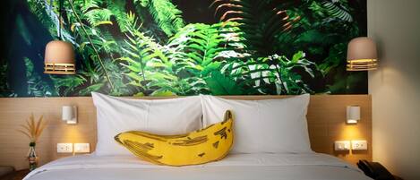 Una habitación de hotel con una cama, un almohadón con forma de plátano, un mural de follaje verde y dos lámparas de pared.