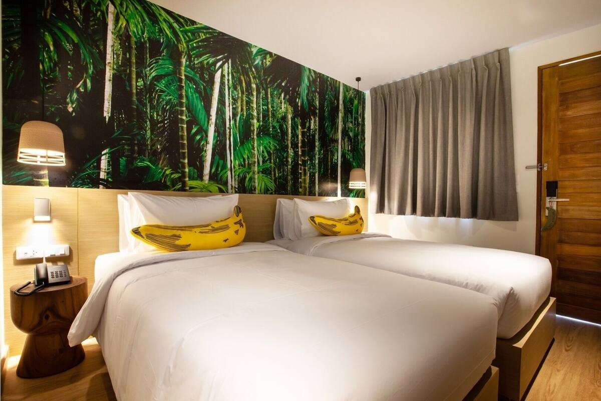 Quarto de hotel com duas camas, um grande mural tropical, uma porta de madeira e um abajur.