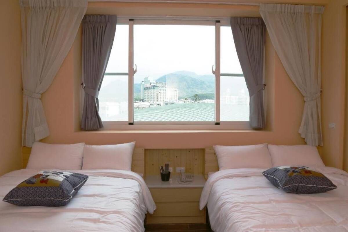 Quarto de hotel com duas camas, uma janela com cortinas, cabeceira de madeira e uma pequena mesa com um telefone e uma xícara.