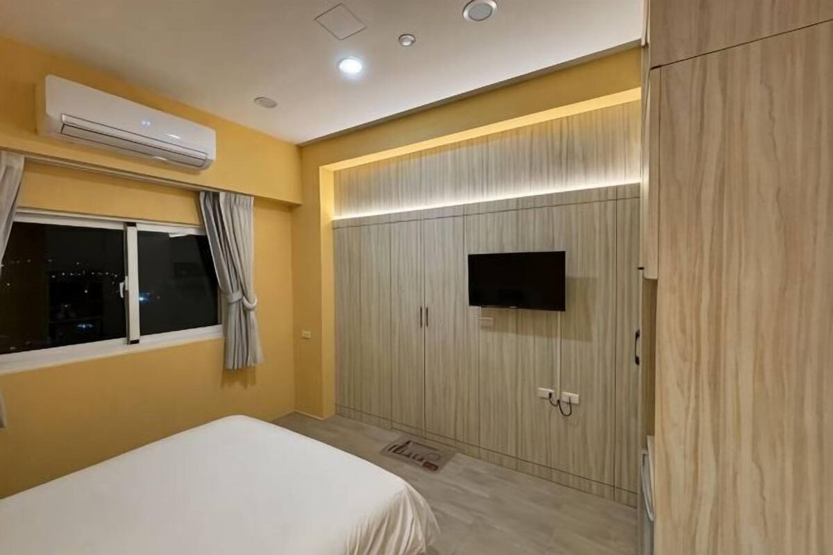 Quarto com cama, ar condicionado, guarda-roupa com televisão e janela com cortinas.