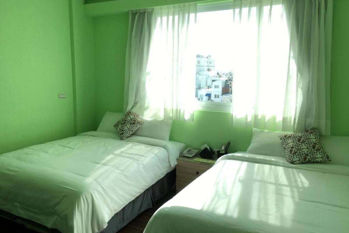 Quarto com duas camas, janela com cortinas e vista para edifícios.