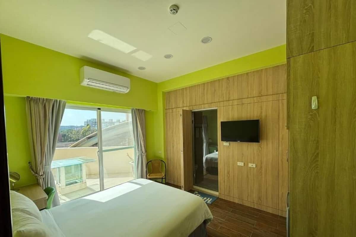Quarto de hotel com cama, televisão na parede, porta deslizante para uma varanda e aparelho de ar condicionado na parede.