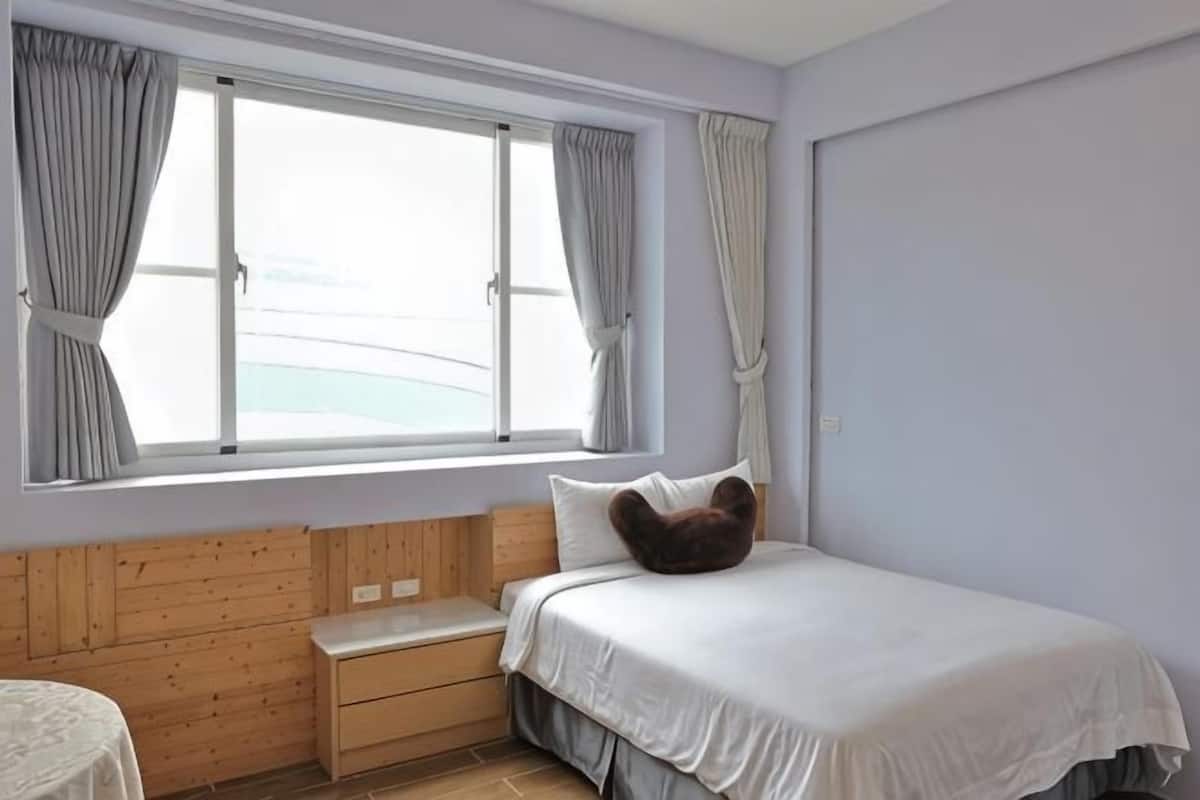 Quarto com uma cama, um banco de madeira, janela com cortinas e vista para o mar.