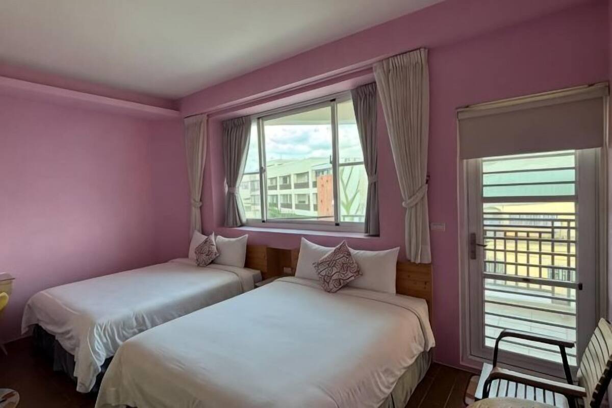 Quarto com duas camas, uma janela com cortinas, uma cadeira e vista para prédios.