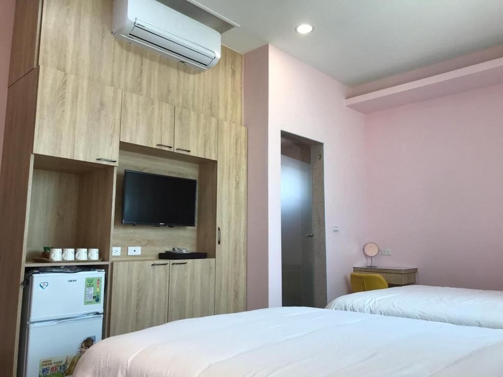 A hotel room with a bed, a TV mounted on a wooden cabinet, a refrigerator, and a small table with a lamp.