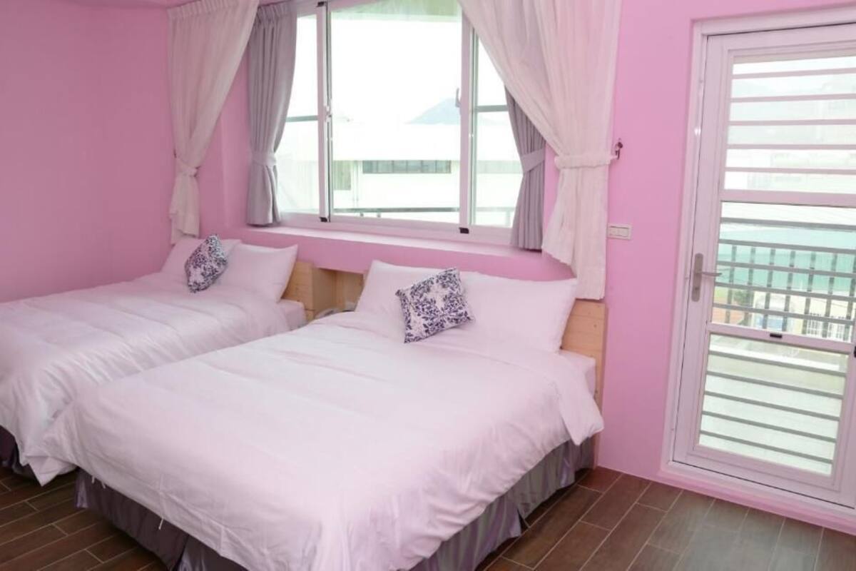 Quarto com decoração em tons de rosa, duas camas, uma janela com cortinas e uma porta de vidro deslizante.