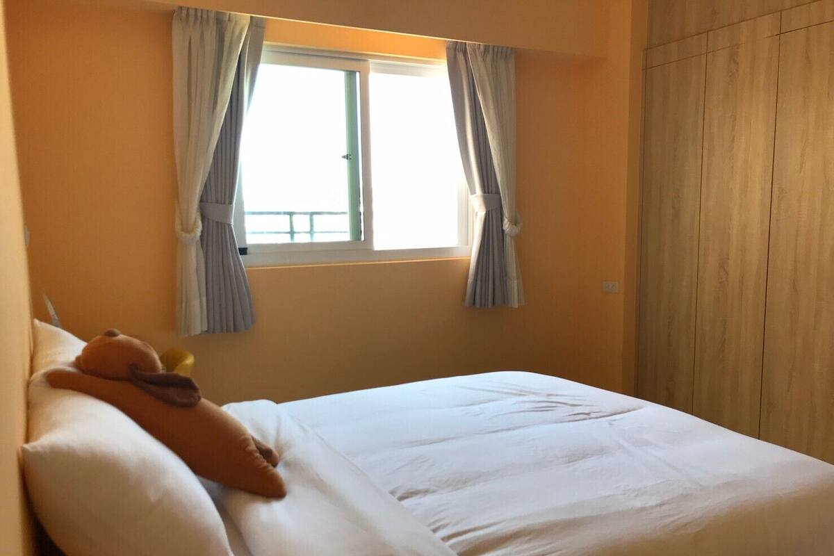 Quarto com cama, almofadas e um ursinho de pelúcia. Há uma janela com cortinas e um guarda-roupa de madeira.