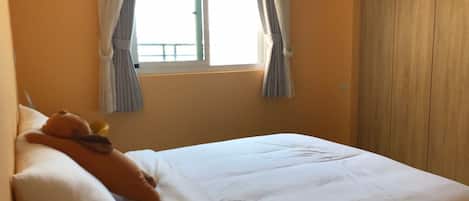 Ein Schlafzimmer mit einem Bett, Kissen und einem Teddybären. Ein Fenster mit Vorhängen und ein Holzschrank sind ebenfalls vorhanden.