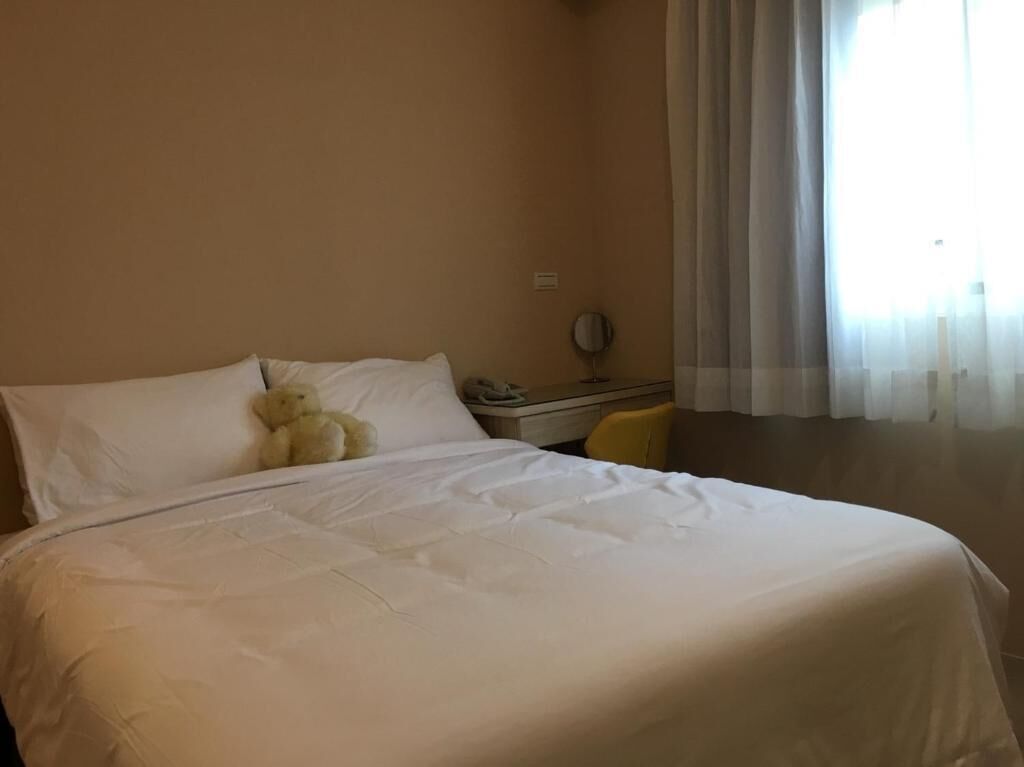Una cama bien hecha con sábanas blancas, una mesita de noche con lámpara y un osito de peluche sobre la almohada.