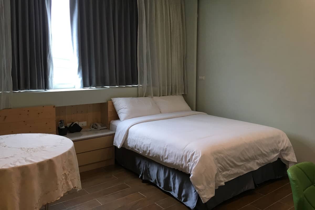 Quarto com cama, mesa de cabeceira, cadeira e janela com cortinas.