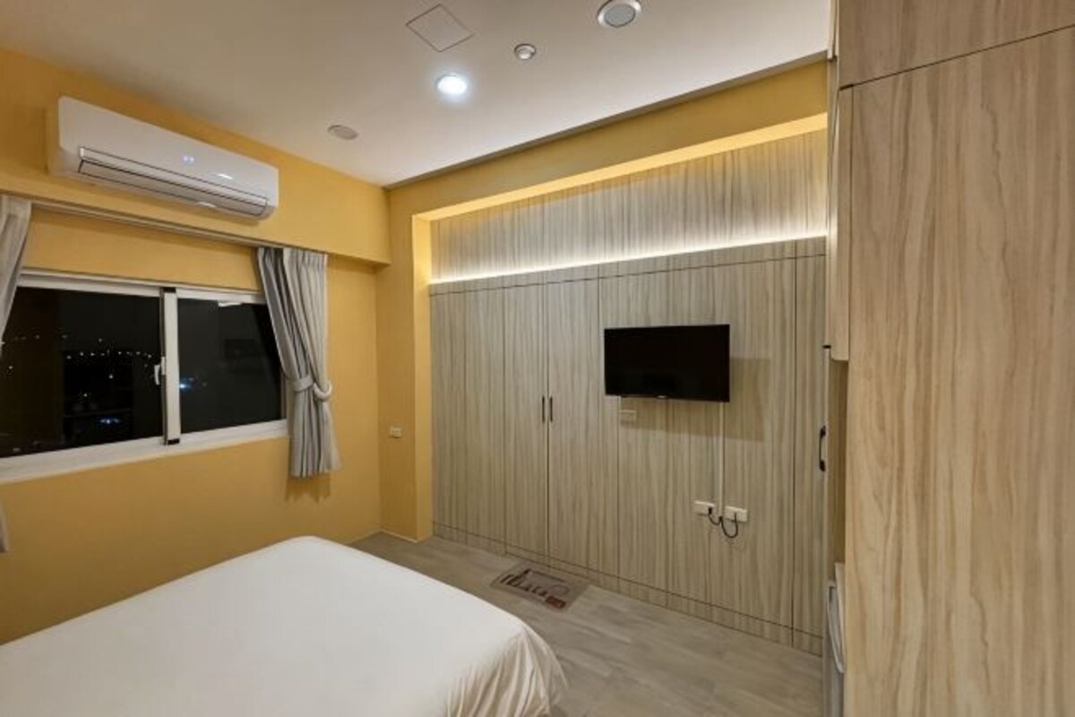 Une chambre à coucher comprenant un lit, un appareil de climatisation, une armoire avec un téléviseur et une fenêtre avec des rideaux.