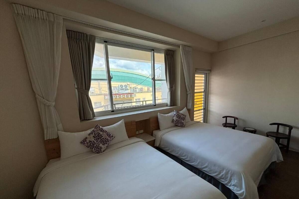 Quarto de hotel com duas camas, uma janela grande com cortinas e vista para prédios.