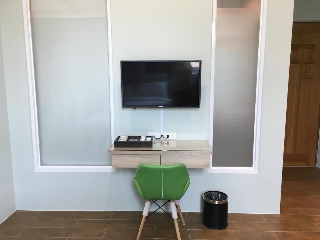 Una habitación moderna con televisión montada, un pequeño escritorio de madera, una silla verde y un cesto de basura.