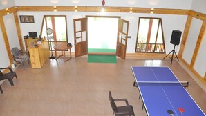 A ping pong table with two paddles, a wooden desk with a computer, a chair, and a green mat in a room with wooden walls and a large window. - Water View Holiday Retreat (Belur)