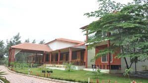 A building with a covered outdoor area, a garden with various plants, and a paved walkway. - Water View Holiday Retreat (Belur)