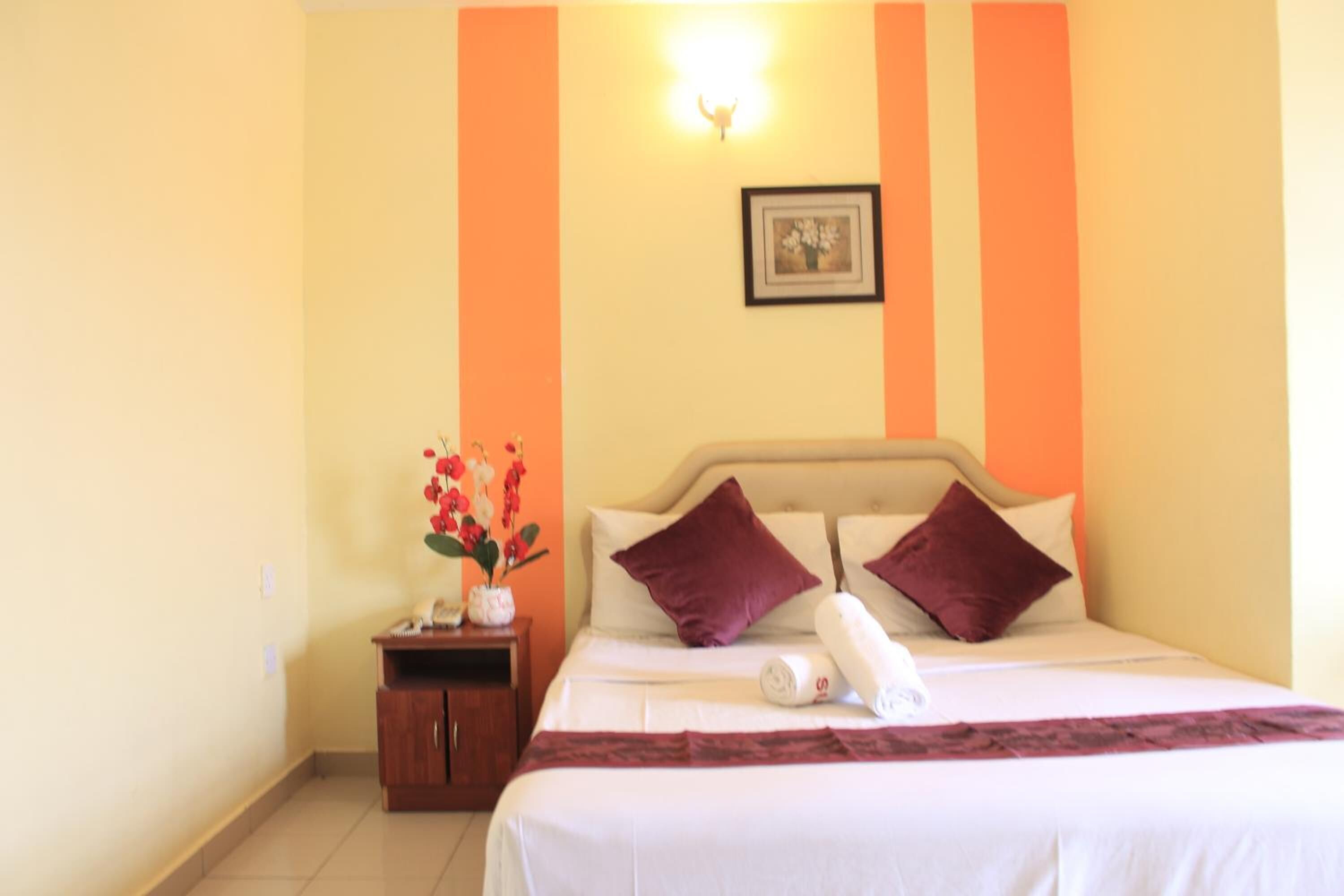 A hotel room with a bed, pillows, a nightstand, a vase with flowers, and a framed picture on the wall.