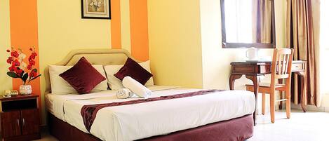 A hotel room with a bed, a desk, a chair, a vase with flowers, and a framed picture on the wall.