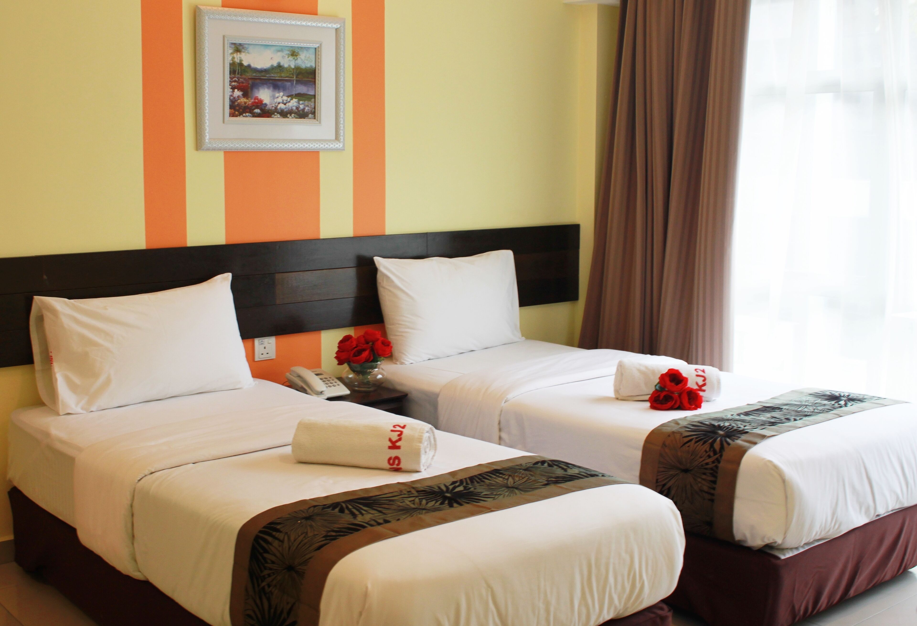 A hotel room with two beds, each with white bedding and a brown headboard. There is a framed picture on the wall above the beds, and a vase with red flowers on the nightstand.