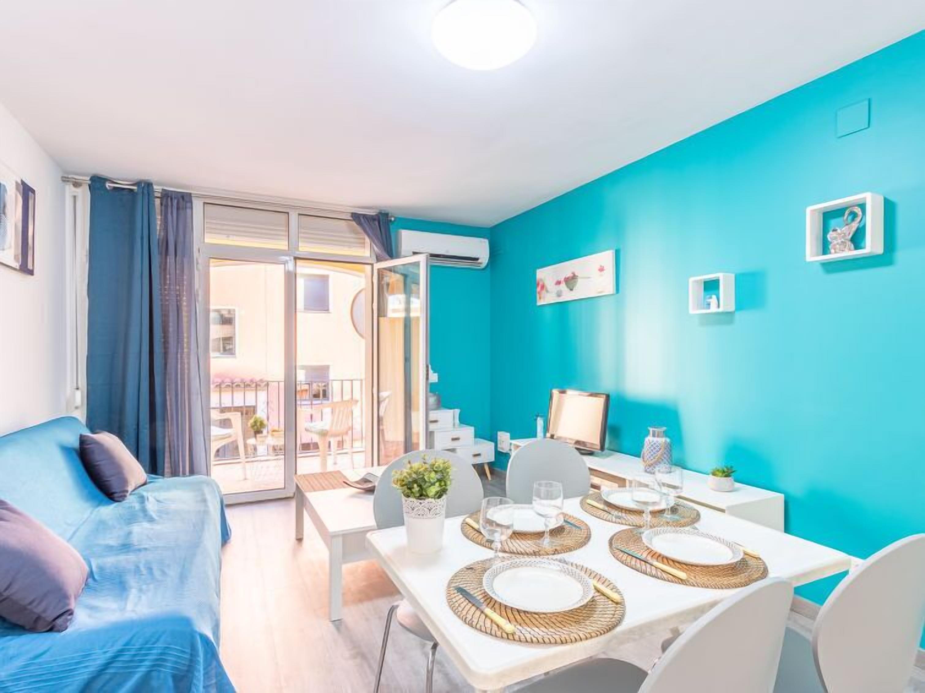 Een lichte kamer met een blauwgroene wand, een eettafel gedekt met borden en glazen, een bank met kussens en een uitzicht door een glazen deur op een buitengebied.