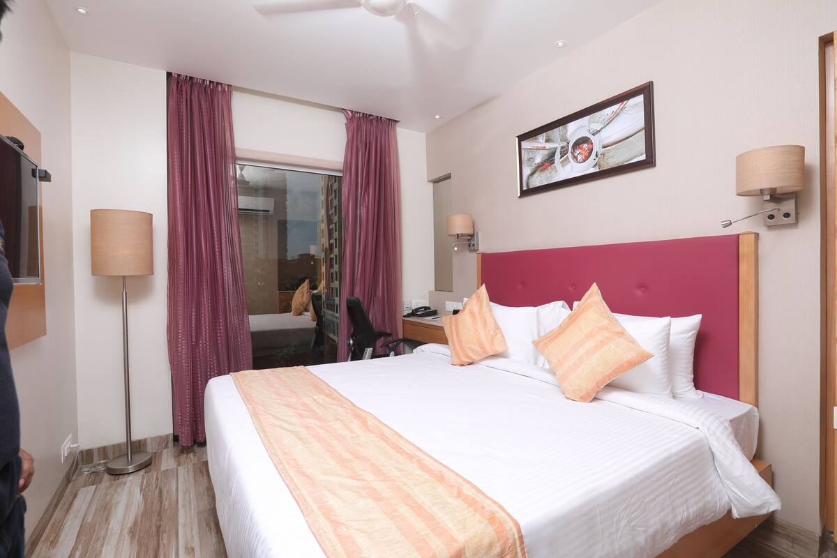 A hotel room with a large bed, a desk with a chair, a standing lamp, and a window with curtains.